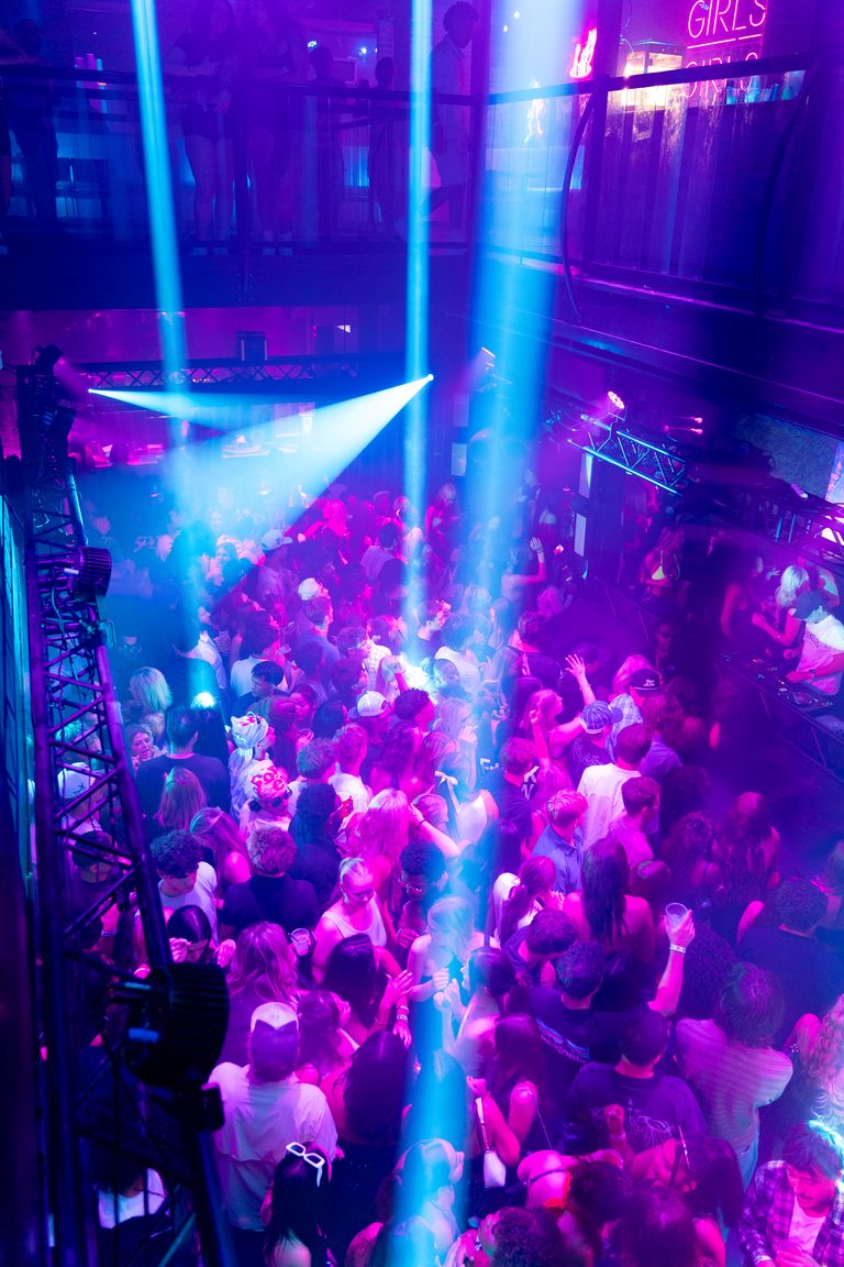 Cyan beams and lasers cut through haze over a packed, magenta-lit dance floor